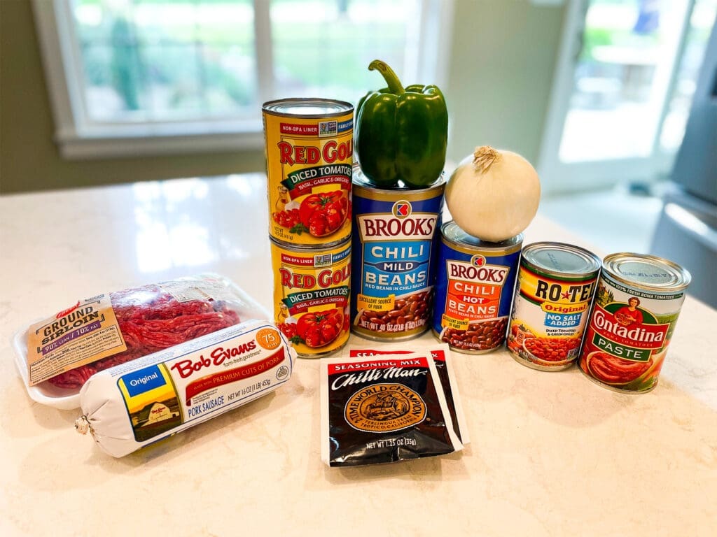 Ingredients for an easy homemade chili recipe displayed on a kitchen counter, including ground sirloin, pork sausage, green bell pepper, onion, Brooks chili beans (mild and hot), Red Gold diced tomatoes, Rotel tomatoes, Contadina tomato paste, and Chilli Man seasoning mix.