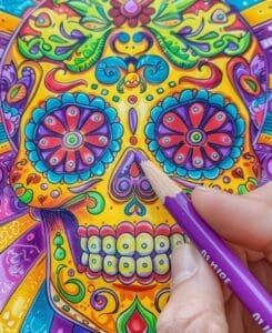 A skull mandala adult coloring page with a hand holding a purple colored pencil, coloring in the page