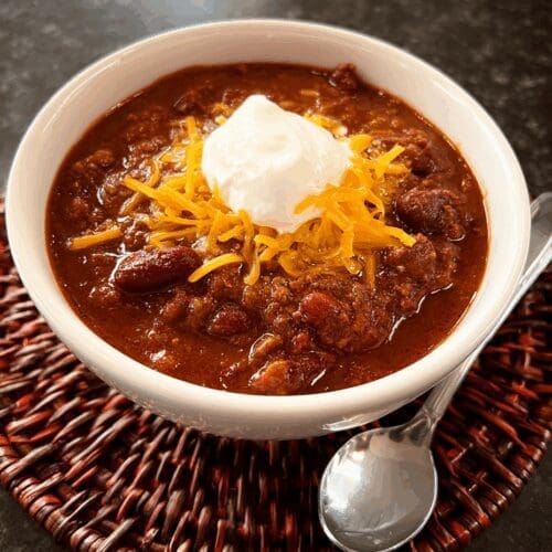 Easy chili recipe served in a white bowl, topped with shredded cheddar cheese and a dollop of sour cream, with a spoon on the side—hearty and perfect for a cozy homemade meal.